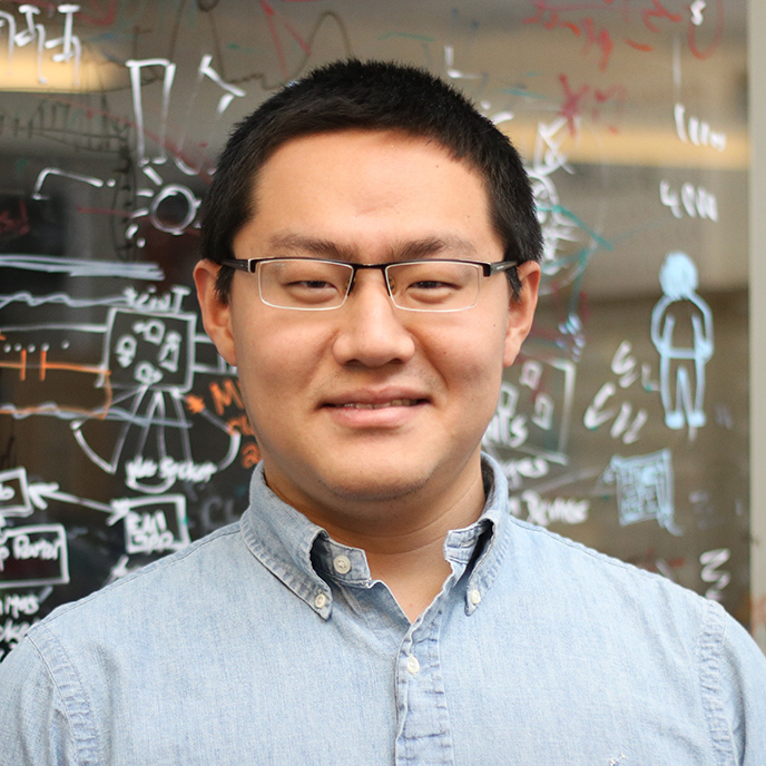 Anhong Guo wearing a light blue shirt standing in front of a glass wall with drawings.