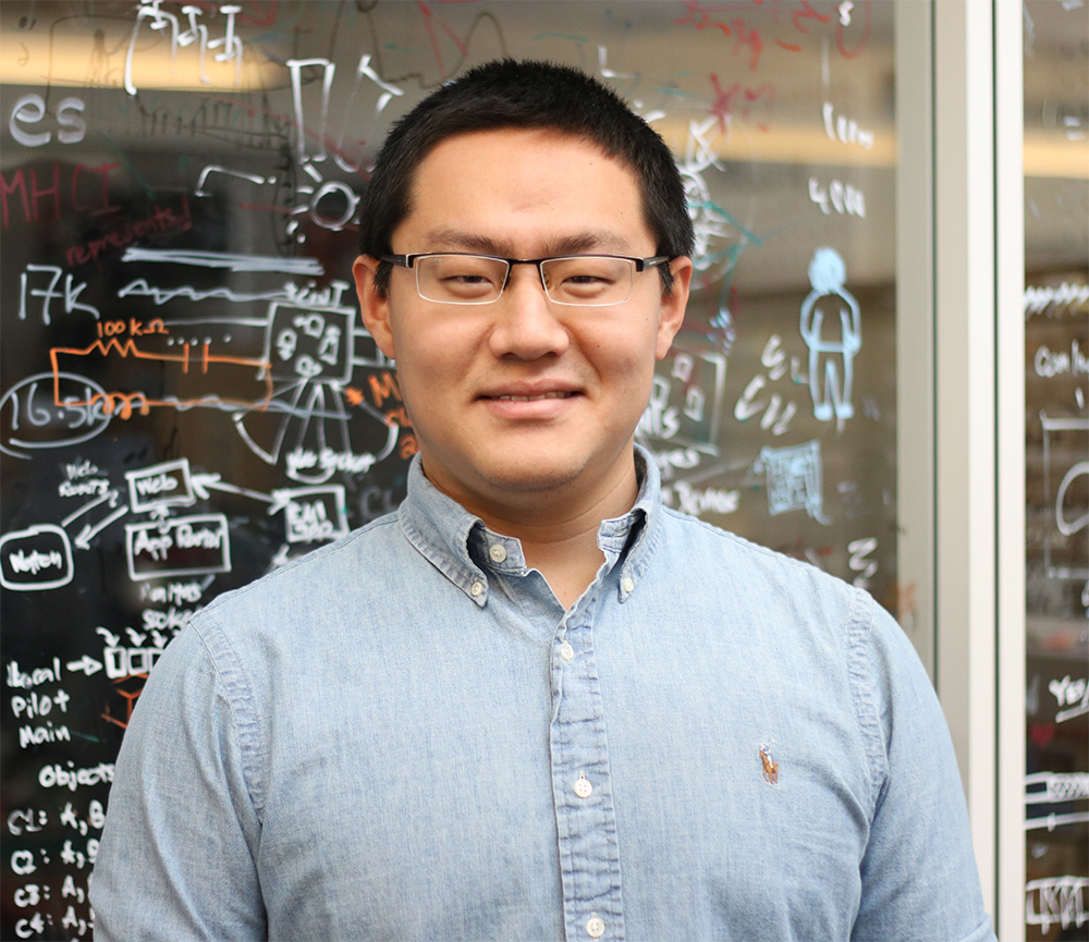 Anhong Guo wearing a light blue shirt standing in front of a glass wall with drawings.