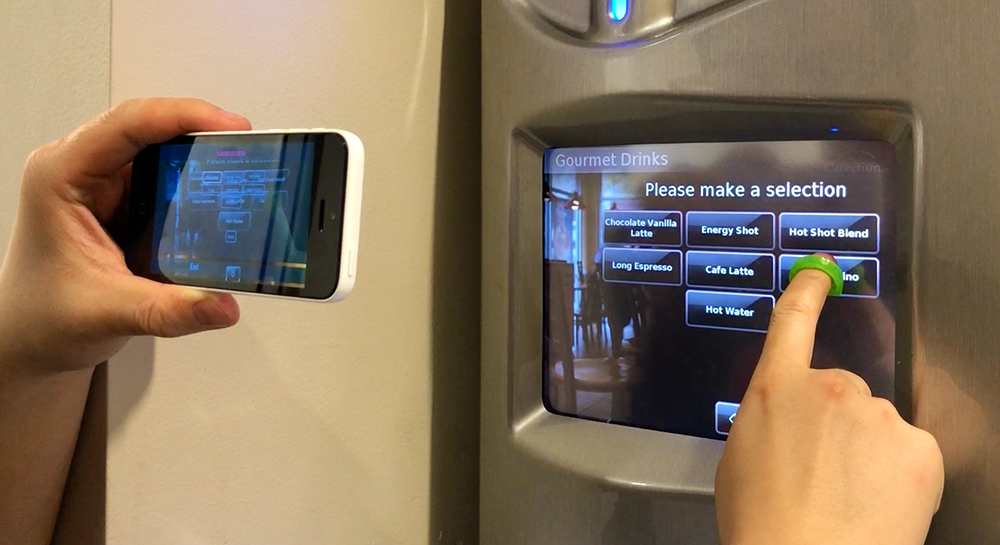 The user is holding the phone in landscape mode with one hand, and aiming the camera towards a touchscreen coffee machine. The user’s other hand is wearing a fingercap exploring on the screen. The StateLens iOS app is providing audio guidance to the user.